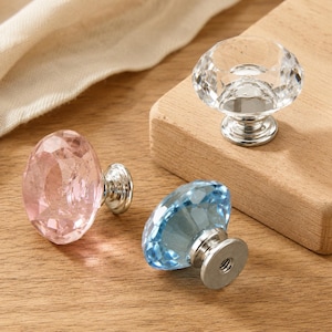 Op de afbeelding: Drie gefacetteerde kastknoppen in diamantvorm, met een glazen bovenkant in transparant, lichtroze en lichtblauw. Elke knop heeft een gepolijste zilverkleurige metalen voet met een zichtbaar schroefgat aan de onderzijde.