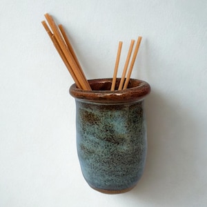 May include: A cylindrical ceramic holder with a mottled blue and brown glaze, featuring a thick, smooth rim. Several thin, light brown wooden sticks are placed inside the vessel, extending upwards from the opening of the container.