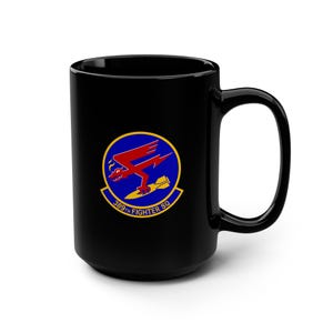 May include: A black ceramic mug featuring a circular emblem of the 389th Fighter Squadron. The logo displays a red winged creature holding a yellow bomb against a blue background, with the text 389TH FIGHTER SQ printed in yellow along the bottom.