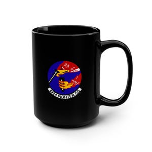 May include: A black ceramic mug featuring a circular emblem of the 492d Fighter Squadron. The logo displays a stylized yellow hand holding a black dagger against a blue and red background, with the text 492d FIGHTER SQ printed in white below.