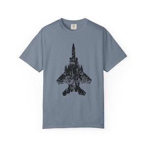 May include: A slate blue short-sleeve t-shirt featuring a large, centered black graphic of a fighter jet. The jet illustration has a distressed, weathered texture. The shirt has a crew neckline and a standard fit, with a small brand label visible at the inner neck.