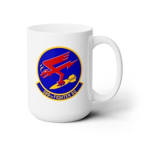 May include: A white ceramic mug featuring the 389th Fighter Squadron emblem. The circular logo displays a red dragon-like creature with a lightning bolt wing, clutching a yellow bomb against a blue background, with the text 389TH FIGHTER SQ.