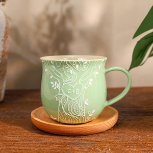 May include: A light green ceramic mug featuring a white line art design of a tree with a heart carved into the trunk containing the letters C+B. The mug has a rounded body, a curved handle, and sits on a circular wooden coaster.