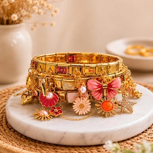 Op de afbeelding: Een goudkleurige modulaire schakelarmband met diverse bungelende bedels, waaronder een roze geëmailleerde strik, een wit madeliefje, een zon met een oranje centrum, een zeester, een schelp met een parel en een klein metalen hartje.