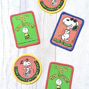 May include: A collection of five embroidered fabric patches featuring Peanuts characters. Two rectangular green patches show Charlie Brown with the text I NEED ALL THE FRIENDS I CAN GET. Two circular pink patches show Snoopy with COME DANCE WITH ME BABY. One rectangular pink patch shows Snoopy as Joe Cool.