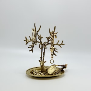 May include: A gold-toned metal jewelry stand featuring a deer figure at the base with branching antlers designed to hold necklaces and rings. The stand includes a circular tray base that holds a wristwatch with a brown leather strap and gold chain.