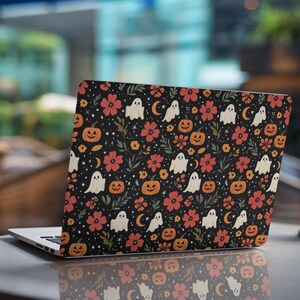 May include: A laptop skin featuring a black background with a repeating pattern of white ghosts, orange jack-o'-lanterns, red and yellow flowers, green leaves, small yellow crescent moons, and tiny white stars covering the entire surface.