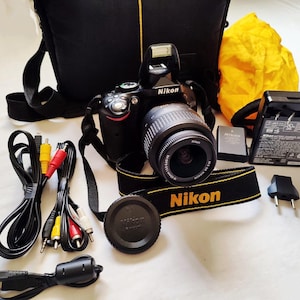 May include: A black Nikon DSLR camera with an attached lens, a black camera bag with yellow trim, a yellow protective pouch, a battery charger, a battery, a lens cap, and various black cables with yellow, red, and white connectors for connectivity.