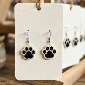 May include: A pair of dangle earrings featuring black and white beaded paw print charms suspended from silver-toned metal hooks. The earrings are displayed on a rectangular off-white card hanging from a wooden stand in a retail setting.