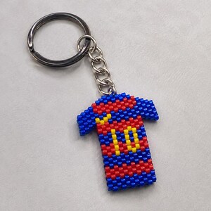 Può includere: Un portachiavi con un ciondolo a forma di maglia in miniatura realizzato con piccole perline quadrate in un motivo a righe orizzontali blu e rosse. Il design della maglia presenta il numero 10 in giallo sul davanti e un piccolo segno giallo.