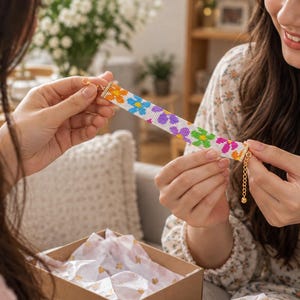 May include: A beaded bracelet featuring a white background with a repeating pattern of orange, blue, purple, and green flowers. The bracelet includes a gold-toned metal clasp and an adjustable chain extension with a small round bead at the end.