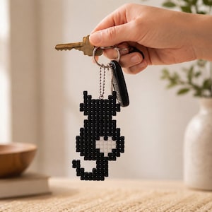 May include: A black cat-shaped keychain constructed from small square beads, featuring a white heart-shaped detail on the side. The keychain is attached to a silver-toned metal ring that also holds a brass-colored key and a black plastic fob.