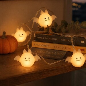 May include: A string of decorative LED lights featuring small, glowing, white bat-shaped covers with simple black eyes and a zigzag mouth. The lights are connected by a thin, flexible wire and emit a warm, soft light, creating a festive atmosphere.