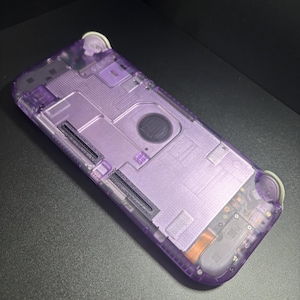 May include: A handheld gaming console featuring a translucent purple plastic shell that reveals the internal components, including metallic plates, circuit board elements, and two light-colored shoulder buttons at the top corners.