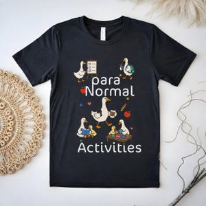Peut inclure: Un t-shirt noir à manches courtes avec un motif représentant cinq oies blanches effectuant diverses activités. Le texte "para Normal Activities" est imprimé en blanc. Les oies sont illustrées avec des objets comme une liste, un sac, un livre et un puzzle.