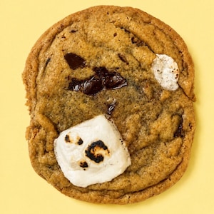 May include: A round, golden-brown baked cookie featuring visible dark chocolate chunks and two pieces of white, toasted marshmallow. The cookie has a textured, slightly uneven surface with a soft, baked appearance and distinct charred spots.