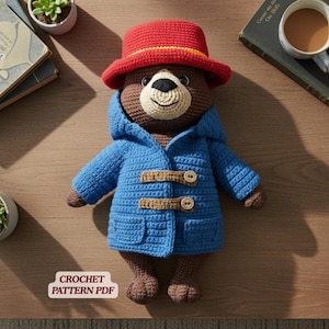 Könnte beinhalten: Eine gehäkelte Bärenpuppe in Braun mit einem leuchtend roten Hut und einem blauen Kapuzen-Dufflecoat mit zwei beigen Knebelverschlüssen. Der Bär hat eine cremefarbene Schnauze und eine schwarze Nase. Ein weißes Etikett mit dem Text CROCHET PATTERN PDF ist sichtbar.