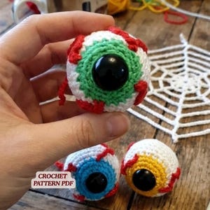 以下が含まれることがあります： 白い糸、赤い刺繍の血管、大きな黒いプラスチックの瞳孔を持つ3つの球形の編みぐるみ眼球。1つは緑色の虹彩、1つは青色、もう1つは黄色の虹彩です。白いラベルにはCROCHET PATTERN PDFと書かれています。