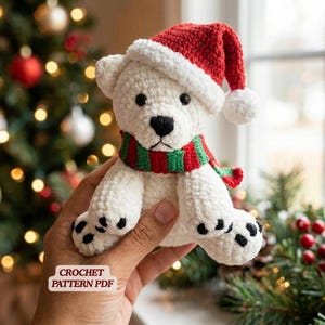 May include: A white crocheted polar bear plush toy wearing a red Santa hat with a white pom-pom and a red and green striped scarf. The bear has black eyes, a black nose, and black paw pads. A white label with the text CROCHET PATTERN PDF is visible.