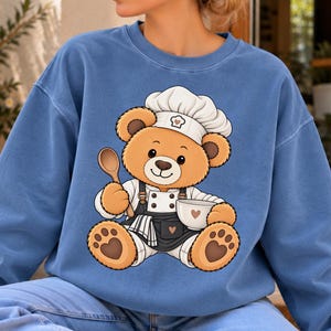 May include: A blue crewneck sweatshirt featuring a graphic of a brown teddy bear dressed as a chef. The bear wears a white chef hat and a black apron, holding a wooden spoon and a white bowl. The bear has heart-shaped paw pads and a small heart on the apron.