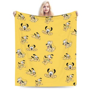 Pode incluir: Uma manta de lã amarela com um padrão repetitivo do cão Pluto em várias poses. A personagem de desenho animado é retratada com as suas orelhas pretas características, pelo castanho claro e coleira verde sobre o tecido amarelo.