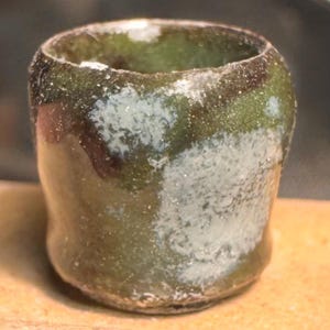 May include: A small, cylindrical ceramic cup featuring a mottled glaze in shades of olive green, dark brown, and patches of white. The surface has a glossy, textured finish with an uneven rim and a slightly tapered base, showing organic color variations.