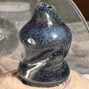 Puede incluir: Un pequeño jarrón de cerámica bulboso con una abertura estrecha en la parte superior. La superficie presenta un esmalte moteado en tonos profundos de azul marino, verde bosque y negro, con un acabado brillante que refleja la luz en su forma.
