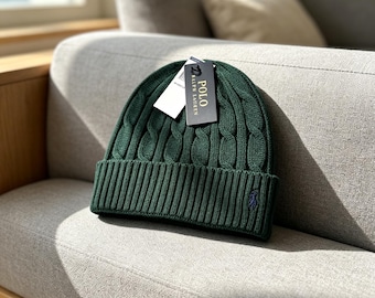 May include: A dark green cable-knit beanie with a wide ribbed cuff. The hat features a small embroidered navy blue logo on the lower right side and a black rectangular tag attached to the top that reads Polo Ralph Lauren in white text.