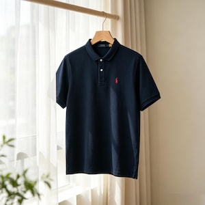 May include: A navy blue short-sleeved polo shirt featuring a classic collar, a two-button placket, and a small red embroidered logo of a person on a horse on the left chest. The garment is displayed on a wooden hanger against a light background.