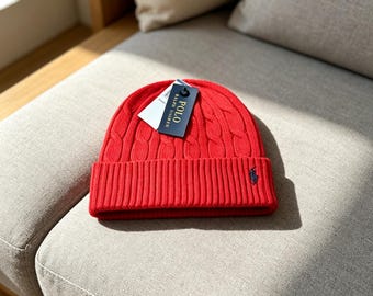May include: A bright red cable-knit beanie with a folded ribbed cuff. The hat features a small navy blue embroidered horse logo on the front cuff and a dark navy rectangular tag attached to the top that reads Polo Ralph Lauren in white text.