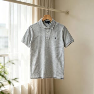 May include: A light gray short-sleeved polo shirt featuring a classic collar, a two-button placket, and a small dark blue embroidered logo on the left chest. The garment is made of a textured knit fabric and is displayed on a wooden hanger.