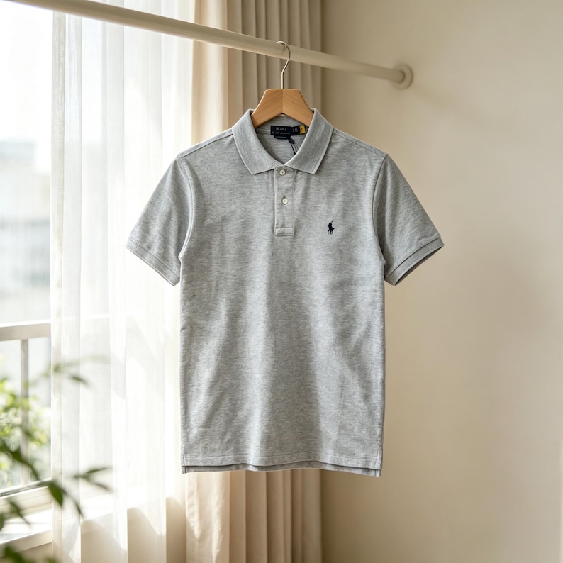 May include: A light gray short-sleeved polo shirt featuring a classic collar, a two-button placket, and a small dark blue embroidered logo on the left chest. The garment is made of a textured knit fabric and is displayed on a wooden hanger.