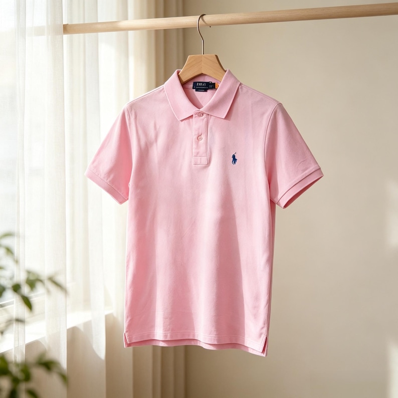 May include: A light pink short-sleeved polo shirt featuring a classic collar, a two-button placket, and a small navy blue embroidered horse logo on the left chest. The shirt is displayed on a wooden hanger against a light, sheer curtain background.