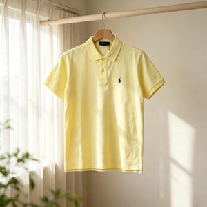 May include: A light yellow short-sleeved polo shirt featuring a classic collar, a two-button placket, and a small navy blue embroidered logo of a person on a horse on the left chest. The shirt is displayed on a wooden hanger against a neutral wall.