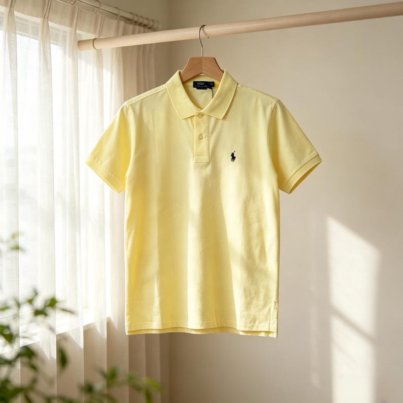 May include: A light yellow short-sleeved polo shirt featuring a classic collar, a two-button placket, and a small navy blue embroidered logo of a person on a horse on the left chest. The shirt is displayed on a wooden hanger against a neutral wall.