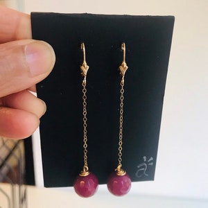 May include: A pair of long, gold chain earrings with burgundy gemstone beads at the end. The earrings are displayed on a black card with the letter 'a' written on it.