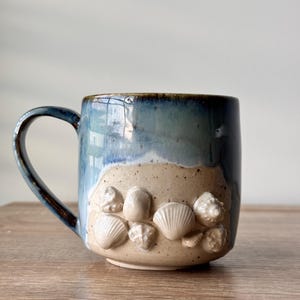 May include: A ceramic mug featuring a two-tone glaze with a deep blue upper section and a speckled beige lower section. The lower portion is decorated with several raised, white, three-dimensional seashell embellishments arranged in a cluster.