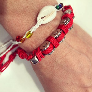 May include: Two bracelets on a wrist. The top bracelet is white with rainbow colored beads and a white seashell charm. The bottom bracelet is red with silver chain links.