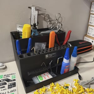 May include: A black plastic Bambu Lab tool organizer with multiple slots and compartments. It holds various tools including a yellow handle, orange scraper, blue and white glue bottles, metal calipers, and scissors. The Bambu Lab logo is printed on the front.