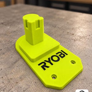 以下が含まれることがあります： ネオンイエローの3Dプリント製バッテリーマウント。平らなベース部分には黒色で「RYOBI」の文字が印字されています。マウントには側面に切り込みが入った中央の垂直タワーと、ベースプレートの前方に2つの円形のネジ穴があります。