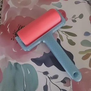 May include: A craft brayer featuring a smooth, cylindrical coral-pink rubber roller attached to a textured, light blue handle. The handle has a small circular hole at the base, and the roller is mounted within a light blue frame that connects to the handle.