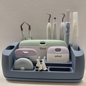 Può includere: Un organizer da scrivania in plastica blu contenente vari strumenti per il fai-da-te, tra cui due raschietti Cricut con manici color lavanda, un grande raschietto Cricut verde, un taglierino circolare, pinzette, un uncino, uno stilo e tappi bianchi.