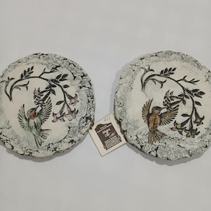 May include: Two circular, textured white ceramic plaques featuring relief carvings of hummingbirds hovering near flowering branches. The left plaque shows a hummingbird with green and red accents, while the right shows a brown hummingbird.