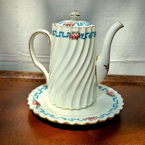 May include: A white porcelain teapot and matching saucer featuring a spiral fluted design. The set is decorated with a blue ribbon pattern and small pink rose clusters, accented with gold trim along the edges, handle, and lid finial.