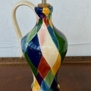May include: A ceramic oil cruet featuring a colorful harlequin diamond pattern in shades of blue, green, yellow, red, and cream. The vessel has a curved handle, a narrow neck, and a cork stopper topped with a silver-toned metal pouring spout.