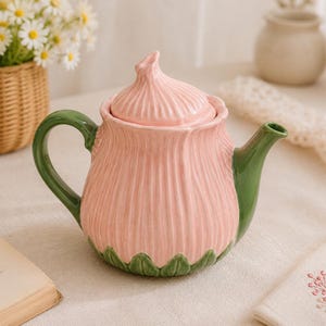 May include: A ceramic teapot shaped like a pink flower bulb with a matching lid. The body features vertical ridges in a soft pink hue, while the handle, spout, and base are finished in a glossy green glaze, resembling the stem and leaves of a plant.