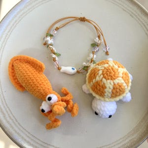 May include: A crochet orange squid, a crochet orange and white turtle, and a bracelet featuring a white fish bead, clear flower-shaped beads, and green leaf accents on a thin brown cord. The items are arranged together on a round ceramic plate.