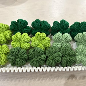 May include: A collection of fifteen crocheted four-leaf clovers in various shades of green, ranging from light lime to deep forest green, arranged on a white rectangular tray with a decorative scalloped edge and a wavy, textured border.