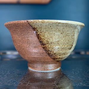 May include: A ceramic bowl featuring a two-tone design with a rough, unglazed reddish-brown clay base on one side and a smooth, speckled cream-colored glaze on the other. The bowl has a wide, flared rim and a small, circular footed base.