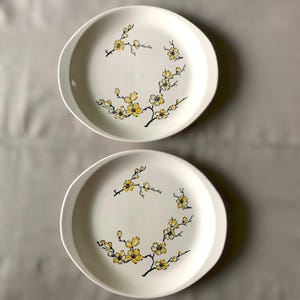 May include: Two round white ceramic plates featuring a delicate hand-painted design of black branches with yellow blossoms. The floral pattern curves along the edge of each plate, creating a circular motif. The plates have a smooth, matte finish.